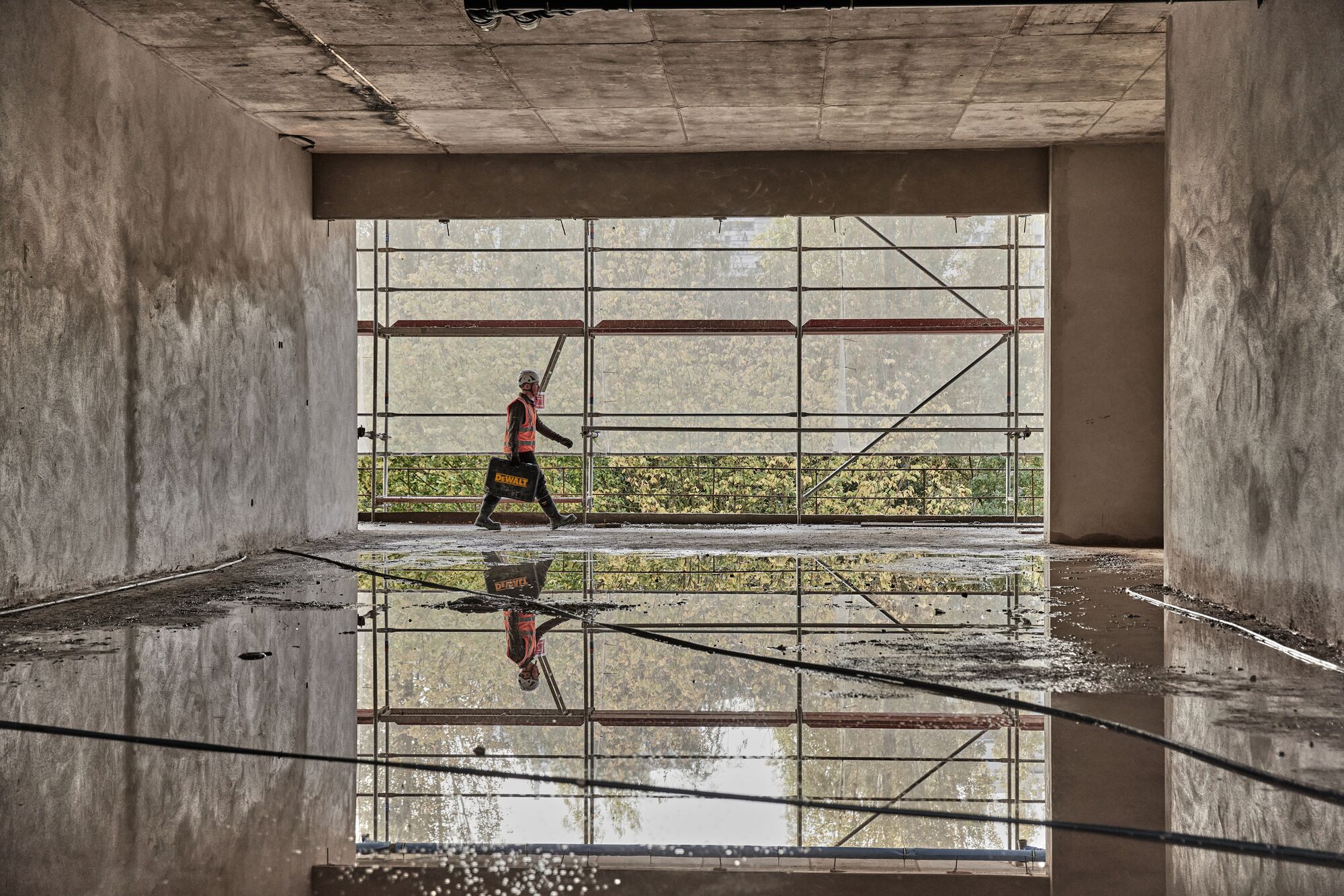 Worker walks with a Dewalt kit case on a construction site. His image is reflected in a puddle of water