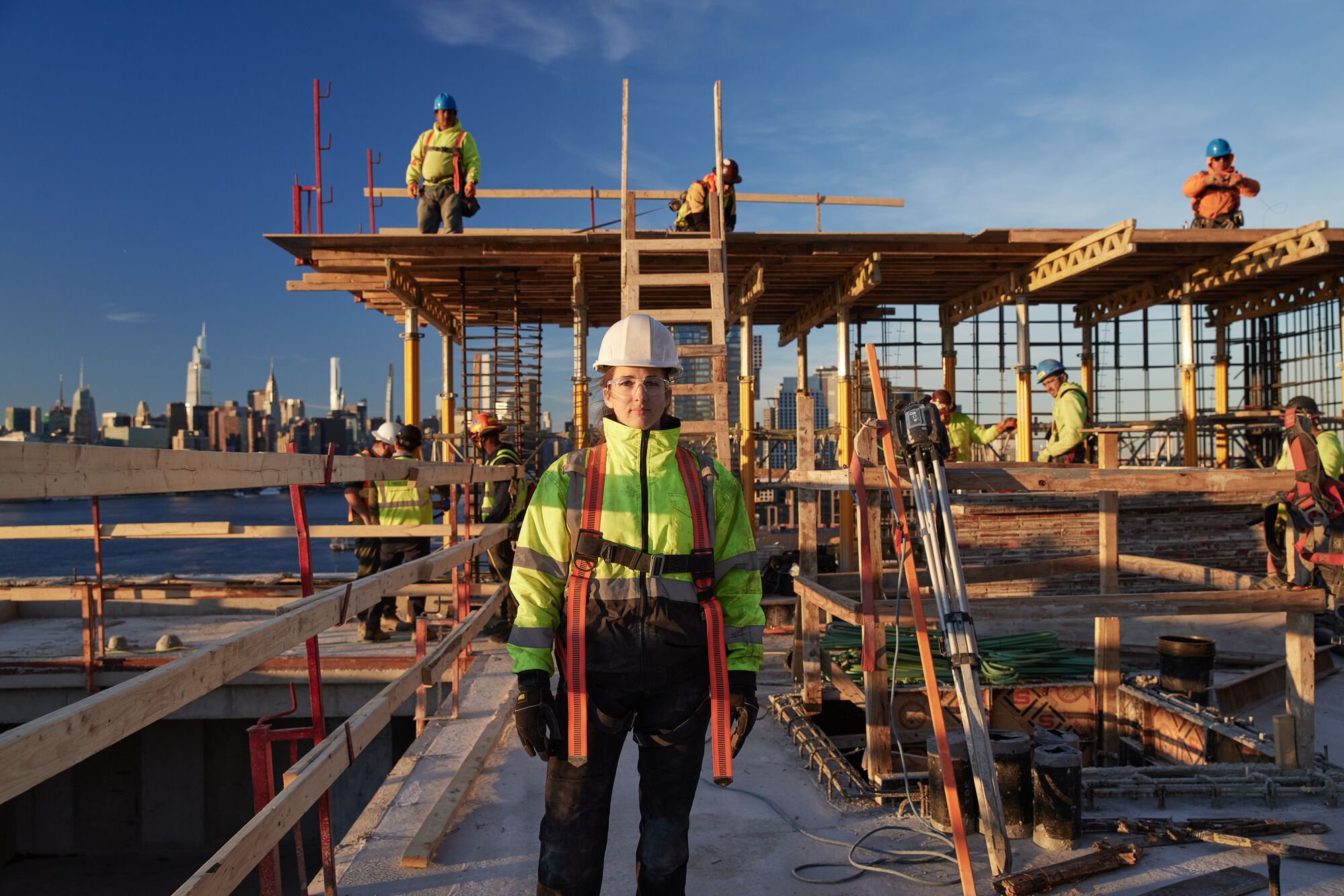 A woman stands at a jobsite looking into the camera. Several tradespeople are working on various tasks