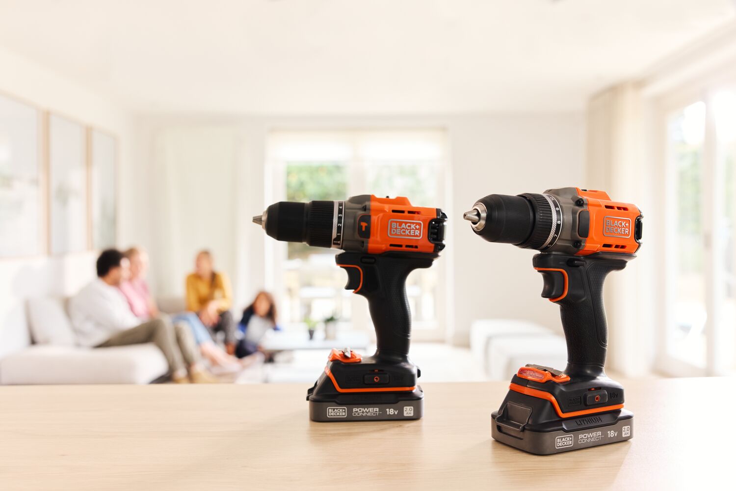 The BLACK+DECKER® POWERCONNECT™ 18V Cordless Hammer Drill and Drill Driver standing on a living room table with a family in the background.