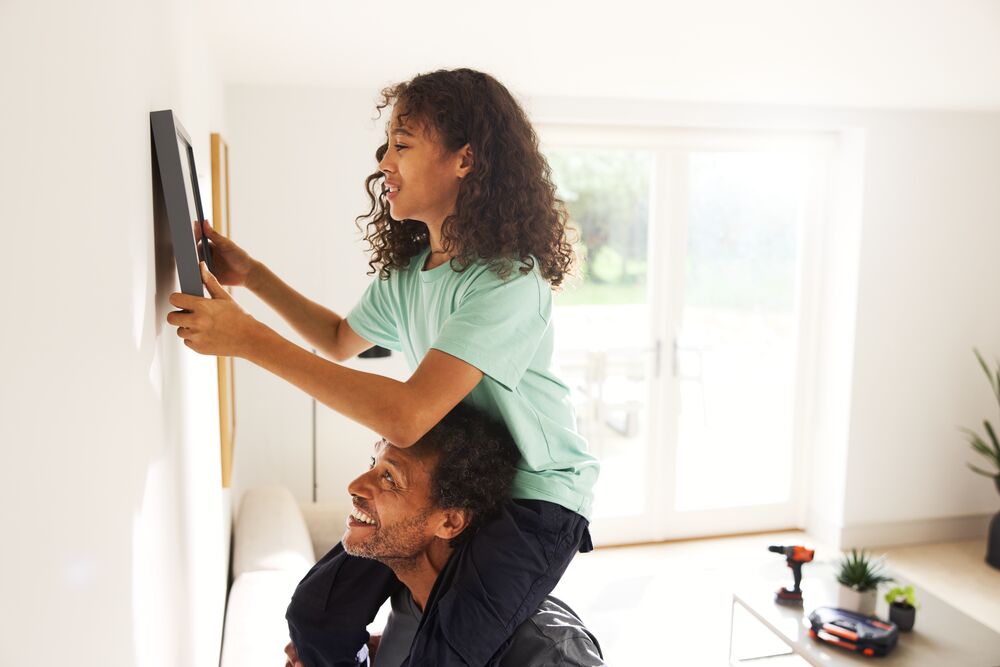 A father and daughter hang a picture frame on a wall with the BLACK+DECKER® POWERCONNECT™ 18V Cordless Hammer Drill in the background.