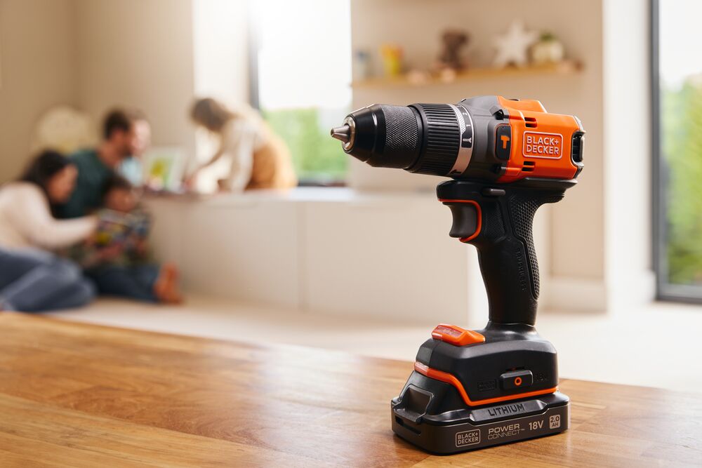 The BLACK+DECKER® 18V POWERCONNECT™ Brushless Hammer Drill standing on a living room table with a family in the background.